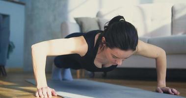 Athletic young woman in black top does push ups near sofa in spacious living room at coronavirus lockdown closeup slow motion video