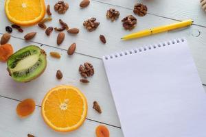 cuaderno con bolígrafo rodeado de manzanas, kiwi, frutos secos, naranjas y manzanas. el concepto de una dieta saludable y una lista de compras. foto