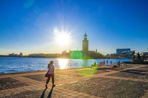 la gente está sentada en un banco en el terraplén del lago malaren mirando el ayuntamiento de estocolmo stadshuset foto