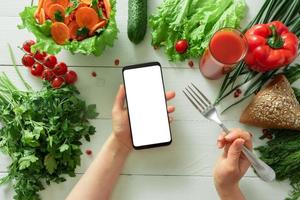 Female hand holds a smartphone on a background of a table with diet vegetables. Place for your text. photo