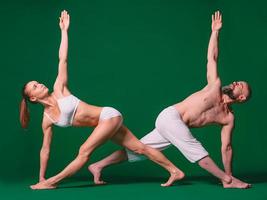Beautiful sporty woman and man in white clothes doing yoga asanas together indoor on green background photo