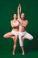 Beautiful sporty woman and man in white clothes doing yoga asanas together indoor on green background photo