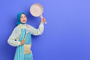 Portrait of smiling young Asian Muslim woman housewife wearing apron holding spatula and frying pan isolated on purple background. Housekeeping concept photo