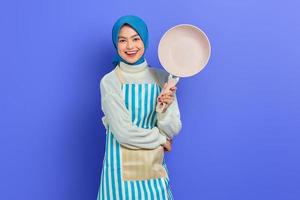 Cheerful beautiful Asian woman in apron and hijab hold pan while doing housework isolated over purple background. Housekeeping concept