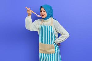 Beautiful Asian woman in white sweater covered in apron and hijab holding spoon, want to taste delicious food isolated over purple background. Housekeeping concept photo