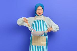 Cheerful beautiful asian muslim woman wearing apron holding frying pan and spatula while doing housework isolated on purple background. Housekeeping concept