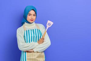 smiling beautiful Asian woman wearing apron and hijab holding spatula isolated over purple background. Housekeeping concept photo