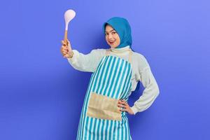 Cheerful young Asian woman housewife in white apron and sweater holding spoon soup while doing housework isolated on purple background studio portrait. housekeeping concept photo