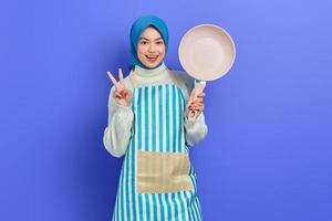 Cheerful beautiful Asian woman in apron and hijab holding pan and showing peace sign with fingers isolated over purple background. Housekeeping concept photo