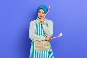 Pensive young Asian woman housewife in white apron and sweater holding spoon soup and spatula while doing housework isolated on purple background studio portrait. housekeeping concept