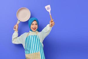 Cheerful beautiful Asian woman in white sweater covered in apron and hijab hold spatula and pan attack enemies while doing housework isolated over purple background. Housekeeping concept photo