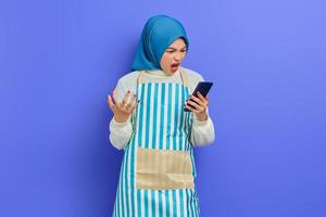 retrato de una joven musulmana asiática enojada que usa hiyab y delantal usando un teléfono móvil mientras grita en voz alta aislada sobre un fondo morado. gente ama de casa concepto de estilo de vida musulmán foto