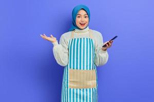 Portrait of excited young Asian muslim woman in 20s wearing hijab and apron using mobile phone while looking at camera isolated over purple background. People housewife muslim lifestyle concept photo