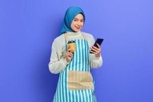 Portrait of smiling young Asian muslim woman in 20s wearing hijab and apron, holding mobile phone and  paper coffee cup isolated over purple background. People housewife muslim lifestyle concept photo