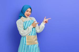 Portrait of smiling housewife woman in hijab and apron pointing product with finger isolated on purple background. People housewife muslim lifestyle concept photo