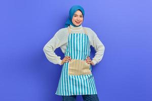 Cheerful young Asian Muslim woman wearing hijab and apron, standing looking at camera with hands on waist isolated on purple background. People housewife muslim lifestyle concept photo