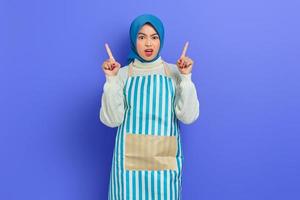 Portrait of displeased housewife woman in hijab and apron pointing at copy space with finger, looking at camera isolated on purple background. People housewife muslim lifestyle concept photo