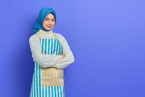 Cheerful young Asian muslim woman wearing hijab and apron crossed arms chest and looking at camera isolated on purple background. People housewife muslim lifestyle concept photo