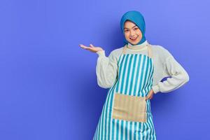 Portrait of smiling housewife woman in hijab and apron showing product with hand isolated on purple background. People housewife muslim lifestyle concept photo