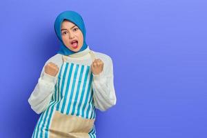 Angry young Asian Muslim woman in hijab and striped apron screaming while clenching hands isolated on purple background.  People housewife muslim lifestyle concept photo