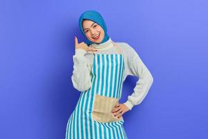 Portrait of cheerful young housewife woman in hijab and striped apron  doing phone gesture like says call me back isolated on purple background. People housewife muslim lifestyle concept photo