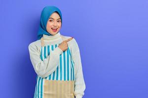 Portrait of excited young housewife woman in hijab and striped apron, standing pointing finger aside on copy space area isolated on purple background. People housewife muslim lifestyle concept photo