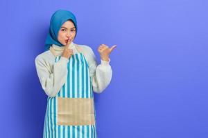 Secret young woman in hijab and stiped apron saying hush be quiet with finger on lips shhh gesture while doing housework, pointing aside with finger isolated on purple background photo