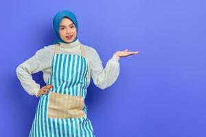 Portrait of smiling young Asian Muslim woman in hijab and striped apron, pointing aside with hands while doing housework isolated on purple background. People housewife muslim lifestyle concept