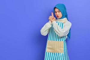 Portrait of serious young housewife woman in hijab and stiped apron,  holding hand like gun gesture isolated on purple background. People housewife muslim lifestyle concept photo