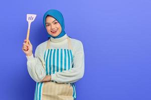 alegre hermosa mujer asiática con suéter blanco cubierto de delantal y hiyab sosteniendo espátula, mirando la cámara haciendo tareas domésticas aisladas sobre fondo morado. concepto de limpieza foto