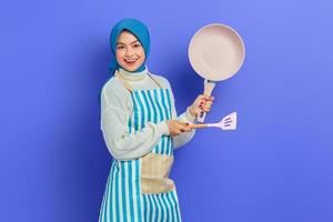 Portrait of cheerful young Asian Muslim woman housewife wearing apron showing frying pan and pointing to copy space with spatula isolated on purple background. Housekeeping concept photo