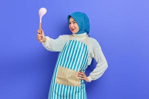 Cheerful young Asian woman housewife in white apron and sweater holding spoon soup while doing housework isolated on purple background studio portrait. housekeeping concept photo