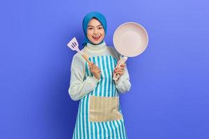 Smiling young Asian woman housewife wearing apron holding frying spatula and pan  while doing housework isolated on purple background. Housekeeping concept photo