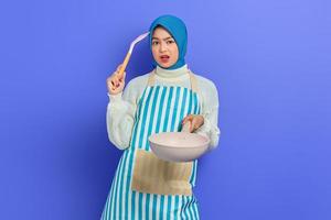 Portrait of confused young Asian woman housewife wearing apron holding frying pan and spatula, looking at camera thinking cooking menu isolated over purple background. Housekeeping concept photo