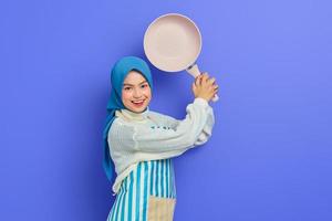 Cheerful beautiful Asian woman in white sweater covered in apron and hijab hold frying pan attack enemies while doing housework isolated over purple background. Housekeeping concept photo