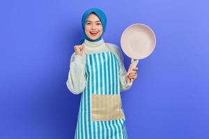 Portrait of excited young Asian Muslim woman wearing apron holding frying pan and celebrating luck isolated over purple background. Housekeeping concept photo