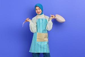 Portrait of smiling young Asian Muslim woman housewife wearing apron holding spatula and frying pan while doing housework isolated on purple background. Housekeeping concept photo
