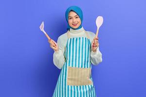 Cheerful beautiful Asian woman housewife in apron holding spatula and spoon soup while doing housework isolated over purple background. Housekeeping concept photo