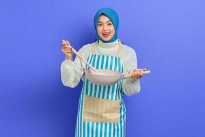 Smiling beautiful Asian woman in white sweater covered in apron and hijab holding spoon and pan while doing homework isolated over purple background. Housekeeping concept photo