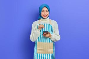 Portrait of excited young Asian Muslim woman in 20s wearing hijab and apron pointing at smartphone with finger isolated on purple background. People housewife muslim lifestyle concept photo