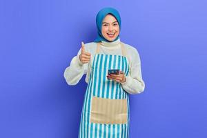 retrato de una joven musulmana asiática sonriente de unos 20 años usando hiyab y delantal usando un teléfono móvil, mostrando un gesto con el pulgar hacia arriba aislado sobre un fondo morado. gente ama de casa concepto de estilo de vida musulmán foto