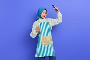 Portrait of excited young Asian muslim woman in 20s wearing hijab and apron holding mobile phone with doing winner gesture isolated on purple background. People housewife muslim lifestyle concept photo