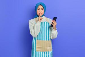 una joven musulmana asiática sonriente que usa hijab y delantal sostiene un teléfono inteligente mientras hace un gesto de silencio aislado en un fondo morado. gente ama de casa concepto de estilo de vida musulmán
