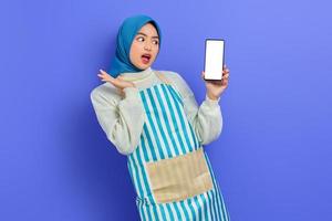 sorprendida joven musulmana asiática usando hijab y delantal sosteniendo un teléfono móvil de pantalla en blanco con la mano aislada en un fondo morado. gente ama de casa concepto de estilo de vida musulmán