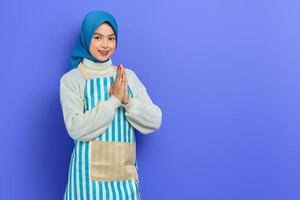 Smiling young Asian Muslim woman in 20s wearing hijab and apron, open sign to greet and greet customers isolated on purple background. People housewife muslim lifestyle concept photo