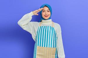 retrato de una joven musulmana asiática emocionada que usa hijab y delantal a rayas, mostrando una actitud lista con las manos en la cabeza mientras hace la tarea aislada en un fondo morado
