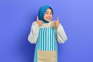 Portrait of cheerful young Asian Muslim woman in hijab and striped apron, standing showing thumbs up and looking camera isolated on purple background. People housewife muslim lifestyle concept photo