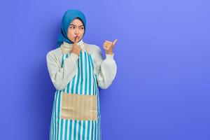 Secret young woman in hijab and stiped apron saying hush be quiet with finger on lips shhh gesture while doing housework, pointing aside with finger isolated on purple background photo