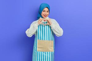 retrato de una joven ama de casa sonriente con hiyab y delantal a rayas, mostrando el corazón en forma con las manos aisladas en un fondo morado. gente ama de casa concepto de estilo de vida musulmán