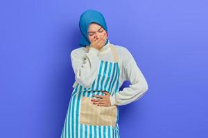 Portrait of sick young housewife woman in hijab and stiped apron,  having abdominal pain, put your hand on your stomach, feel nauseous and want to throw up isolated on purple background photo